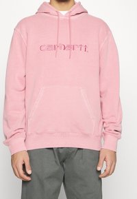 Růžová bavlněná mikina s kapucí, přední kapsou, stahovací šňůrkou a vyšitým logem "Carhartt" ve stejné barvě na hrudi.
