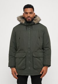 Parka verde con capucha forrada de piel, cierre de cremallera, dos bolsillos delanteros con solapa y cordones ajustables. La tela tiene una textura suave.