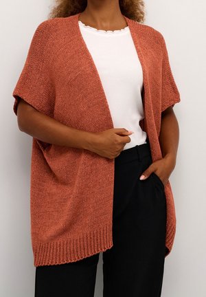 Femme portant un cardigan ouvert tricoté couleur rouille sur un haut blanc à bord festonné et un pantalon noir, posant avec une main dans la poche.
