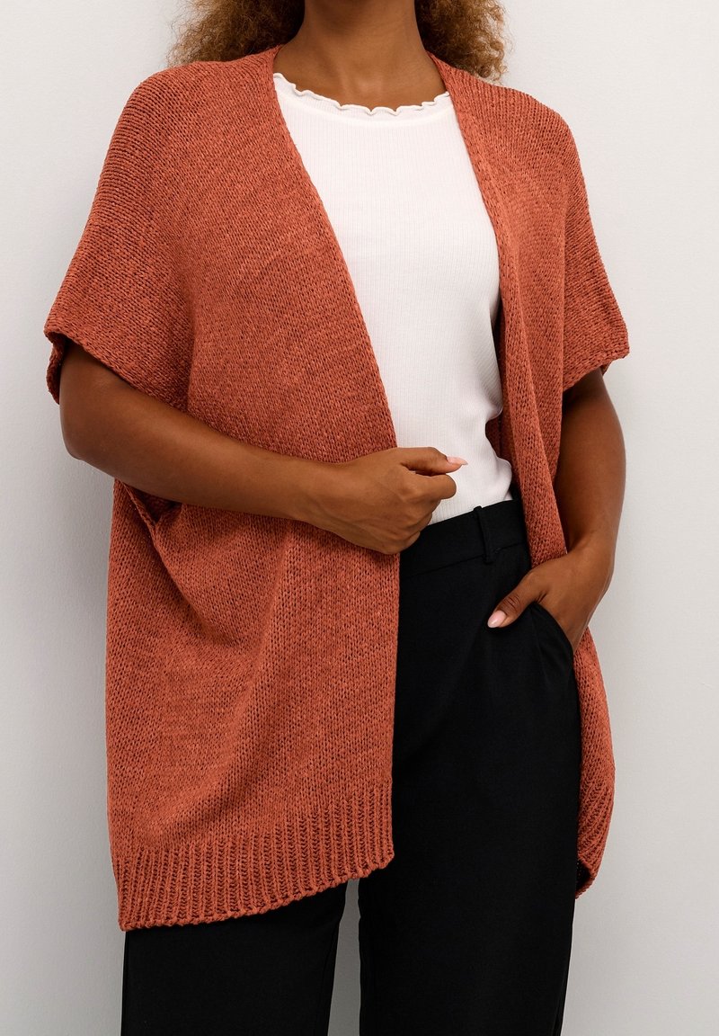 Femme portant un cardigan ouvert tricoté couleur rouille sur un haut blanc à bord festonné et un pantalon noir, posant avec une main dans la poche.