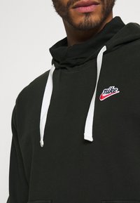 Green hoodie with a high neck design, white drawstrings, and a small embroidered Nike logo in red and blue on the chest. Smooth texture.