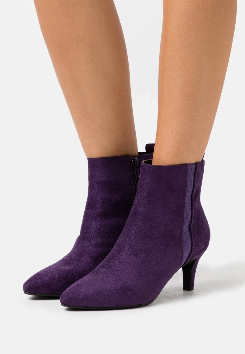 Jambes portant des bottines en daim violet à bouts pointus et talons bas sur fond blanc.