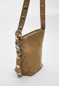 Borsa a mano in suede marrone con base quadrata, chiusura con zip e tracolla perforata con accenti circolari e anelli in argento.