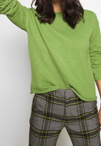 Groene trui met textuur en lange mouwen, gecombineerd met grijze geruite broek met gele accenten en een losse pasvorm bij de taille.