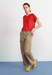 Rotes kurzärmliges Oberteil mit weißen Akzenten und V-Ausschnitt, kombiniert mit beigefarbenen Cargo-Hosen mit großen Seitentaschen. Gelbe Schuhe runden den Look ab.