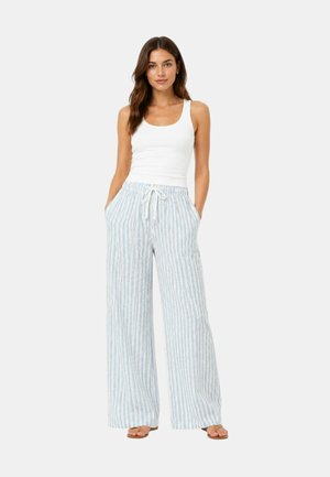 Donna in piedi, indossa una canottiera bianca e pantaloni ampi a righe azzurri con vita regolabile con coulisse, mani nelle tasche, sfondo neutro.