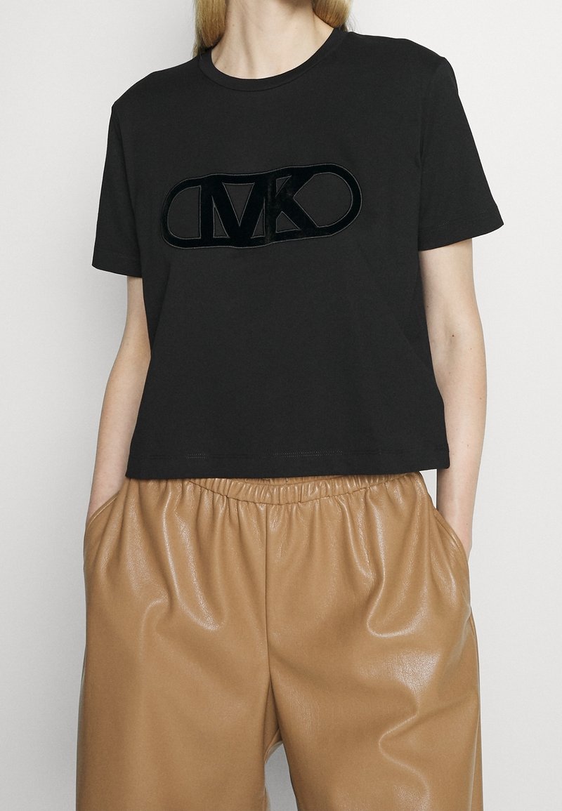 Camiseta negra corta con un logo "MK" negro brillante en el frente, combinada con pantalones beige brillantes de cintura elástica.