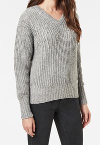 Pull en maille gris avec col en V, présentant une texture côtelée, des manches longues et un ourlet légèrement plus long à l'arrière. Confectionné en tissu doux.