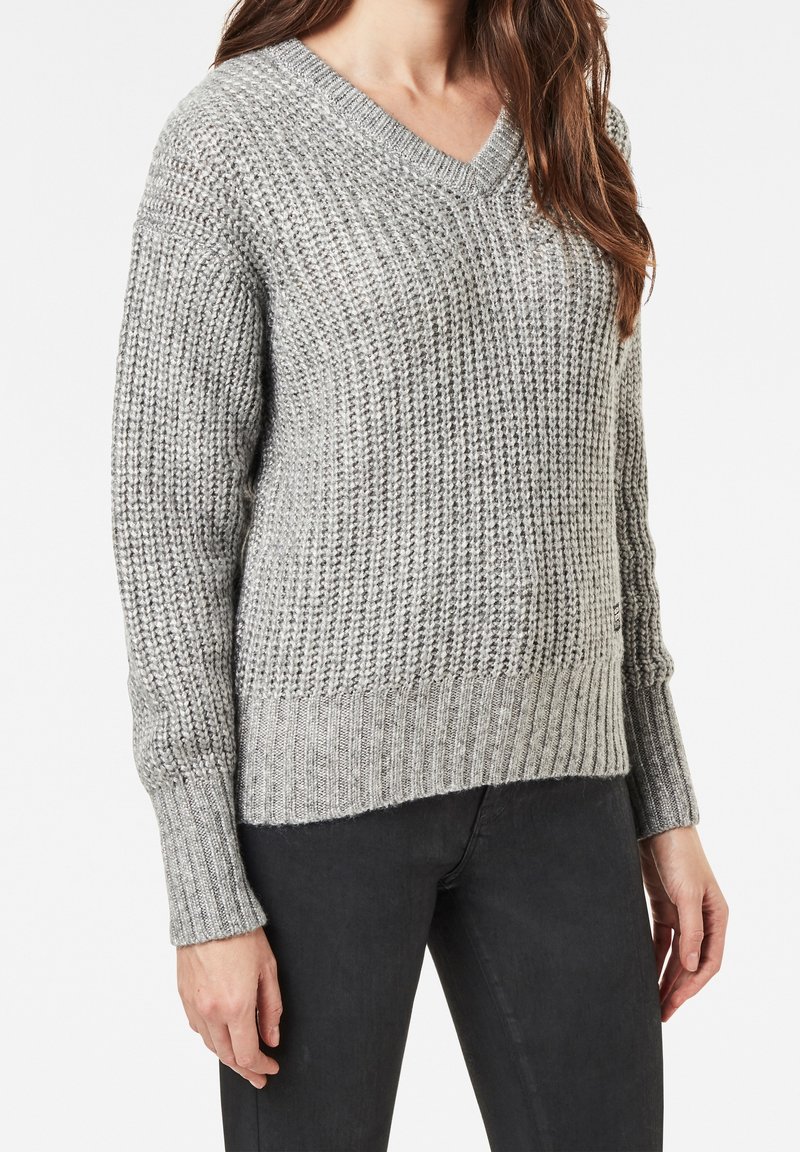 Pull en maille gris avec col en V, présentant une texture côtelée, des manches longues et un ourlet légèrement plus long à l'arrière. Confectionné en tissu doux.
