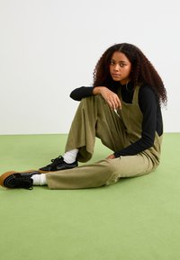 Salopette in corda verde oliva con gambe larghe, abbinata a una maglietta nera a maniche lunghe, sneakers nere e calzini bianchi alla caviglia su un pavimento verde.