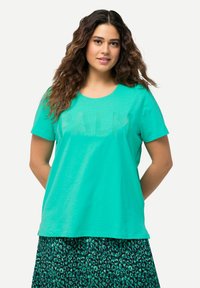 Camiseta corta de manga corta en turquesa con lettering texturizado "PALM" en la parte frontal, escote redondo, hecha de tela suave, llevada sobre una falda de estampado de leopardo verde.