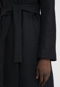Cappotto in lana nera con cintura in vita, caratterizzato da un tessuto testurizzato, maniche lunghe e tasche laterali.
