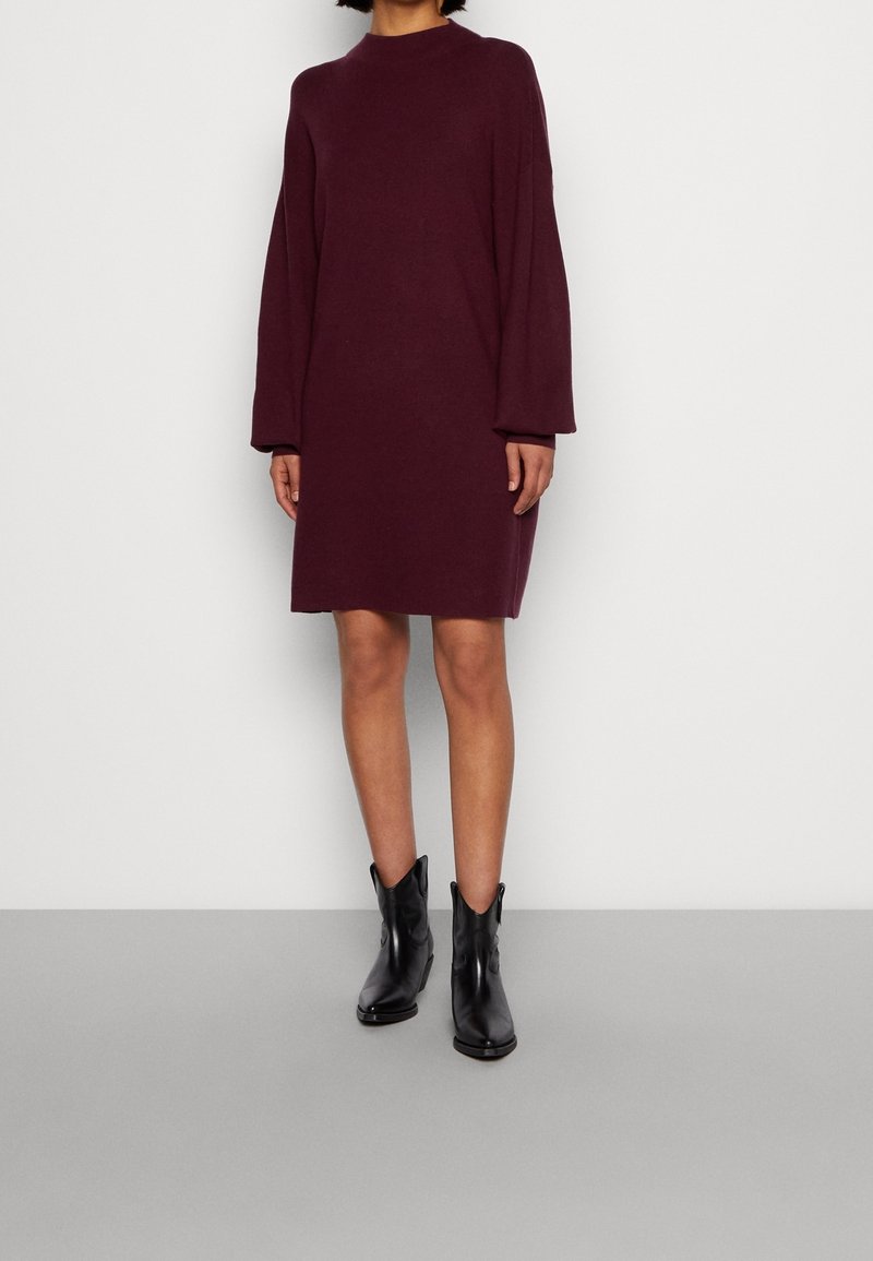 Knielanges, kastanienbraunes Strickkleid mit langen Ärmeln und hohem Kragen. Getragen mit schwarzen Leder-Stiefeletten auf einem grauen Hintergrund.