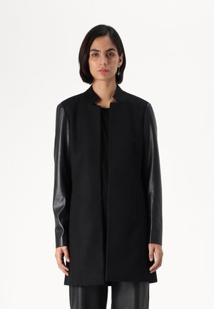 Manteau noir avec un design à col, en tissu de laine et manches en cuir. Le manteau est droit, atteignant le milieu de la cuisse, sans fermetures visibles.