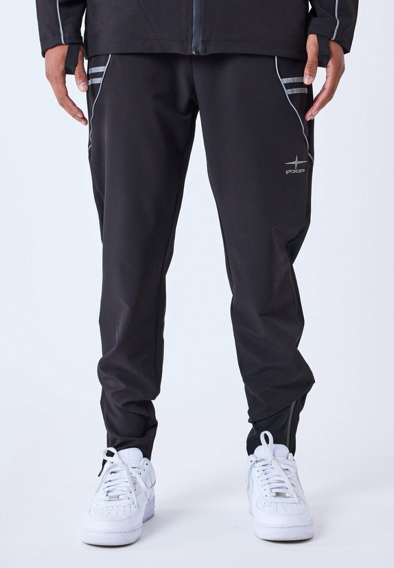 Pantalones deportivos negros de corte ajustado, con bolsillos laterales, detalles reflectantes y puños elásticos, combinados con zapatillas blancas.