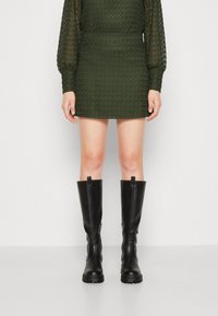 Botas negras hasta la rodilla hechas de material suave con suela gruesa y cremallera lateral, combinadas con un vestido mini verde texturizado.