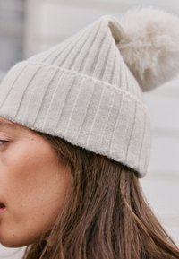 Berretto lavorato a maglia grigio chiaro con trama a coste verticali, caratterizzato da un soffice pon pon sulla sommità. Vista ravvicinata con capelli parzialmente visibili.