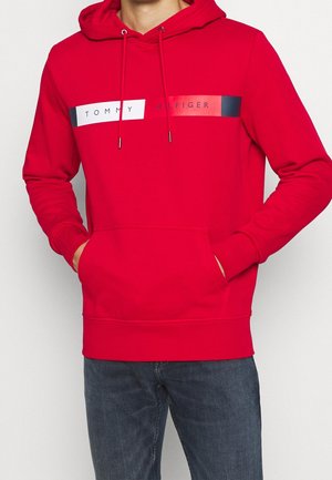 Czerwona bluza z kapturem, przednią kieszenią i sznurkami, z poziomymi białymi, czerwonymi i czarnymi paskami oraz napisem "Tommy Hilfiger" na piersi.