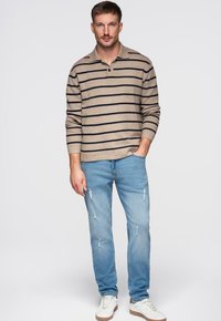 Polo beige à manches longues avec des rayures bleu marine, associé à un jean bleu clair déchiré et des baskets blanches à semelle marron.