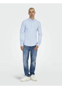 Chemise boutonnée à manches longues bleu clair associée à un jean bleu et des baskets blanches. L'arrière-plan est simple et minimaliste.