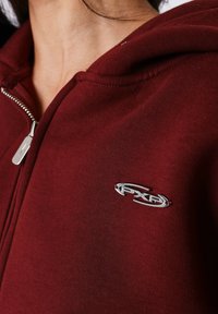 Sudadera con cremallera color burdeos hecha de tela suave, con un acento de logo plateado en el pecho y capucha con cordón.