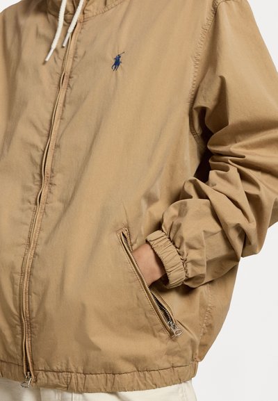 Tan spalvota zip-up striukė su elastinėmis rankovėmis ir raišteliu ant apykaklės. Ant krūtinės yra mažas tamsiai mėlynas logotipas, o šoninėse zip kišenėse.