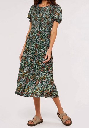 Femme portant une robe midi verte à fleurs avec des manches bouffantes et des sandales beige à bout ouvert, debout devant un fond uni.