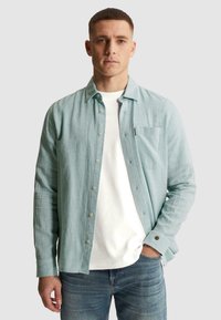 Jeune homme portant une chemise bleu clair boutonnée sur un t-shirt blanc et un jean bleu, debout avec une main dans la poche devant un fond uni.