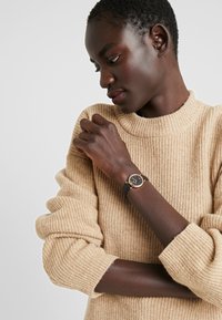 Schwarze Armbanduhr mit einem runden, goldfarbenen Gehäuse, schwarzem Zifferblatt und schwarzem Lederarmband, getragen am Handgelenk einer Person mit einem beigen gerippten Pullover.