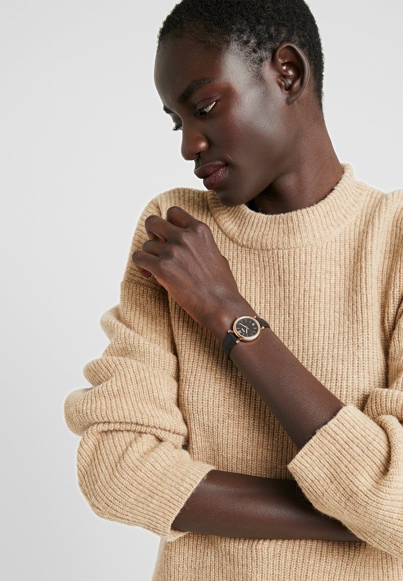 Schwarze Armbanduhr mit einem runden, goldfarbenen Gehäuse, schwarzem Zifferblatt und schwarzem Lederarmband, getragen am Handgelenk einer Person mit einem beigen gerippten Pullover.