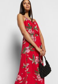 Vestido longo floral vermelho com alças finas, apresentando flores em tons de rosa e verde. Modelo segura uma bolsa de palha preta. Tecido suave, corte descontraído.