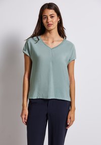 T-shirt verde chiaro a V, con maniche corte arrotolate, realizzato in tessuto morbido, indossato con pantaloni blu scuro; taglio casual e rilassato.