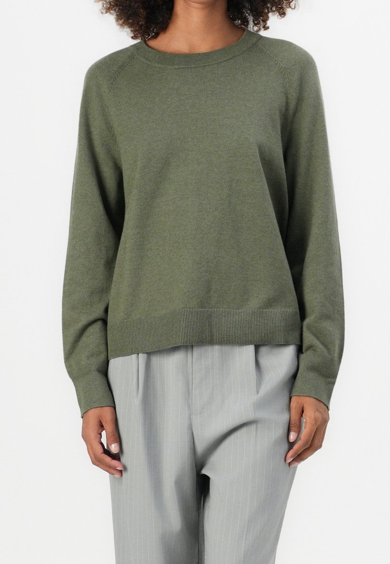 Femme portant un pull vert olive à manches longues et col rond, associé à un pantalon plissé gris clair à rayures fines, sur fond blanc.