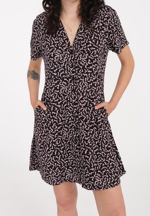 Woman wearing a short-sleeve black dress with pink abstract squiggle patterns, front buttons, and hands in pockets.