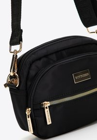 Czarna nylonowa torba na ramię z złotymi akcentami suwaka i logo. Posiada dwie przegrody i regulowany pasek. Gładka tekstura.
