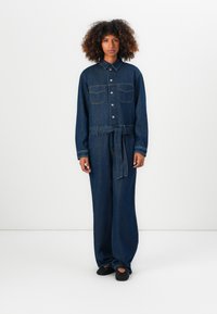Vrouw met krullend haar die een donkerblauwe spijkerbroek jumpsuit draagt met een knoopsluiting aan de voorkant, borstzakken, een tailleband en zwarte geperforeerde schoenen, staand op een witte vloer.