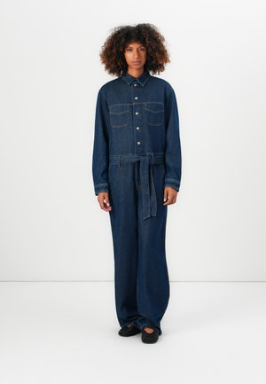Vrouw met krullend haar die een donkerblauwe spijkerbroek jumpsuit draagt met een knoopsluiting aan de voorkant, borstzakken, een tailleband en zwarte geperforeerde schoenen, staand op een witte vloer.