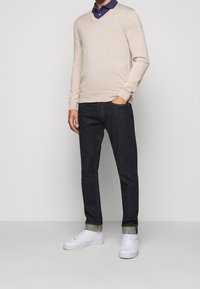 Beige v-ringad tröja i lätt tyg, kombinerad med mörkblå denimjeans och vita sneakers. Tröjan har en subtil textur och logotyp.