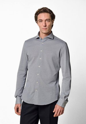 Uomo che indossa una camicia azzurra chiara con bottoni a maniche lunghe e pantaloni neri, in piedi contro uno sfondo bianco e uniforme.