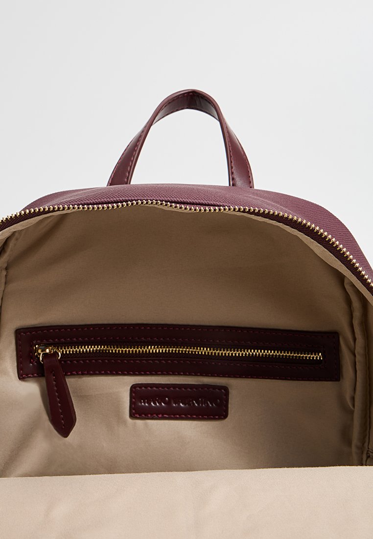 Burgunderfarget lærsekk med gullglidelås, har et beige stoffinteriør, en glidelommer og en etikett i interiøret.