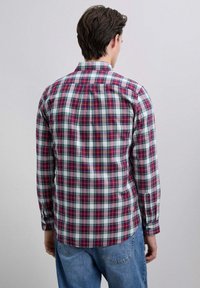 Uomo che indossa una camicia a maniche lunghe a quadri rossi, bianchi e verdi e jeans blu, visto di spalle su uno sfondo grigio chiaro.