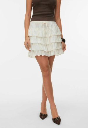 Femme portant une mini-jupe à volants crème à étages avec une taille à nouer, un top marron sans manches, un bracelet noir et des talons marron à bout pointu.