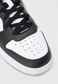 Sneaker noir et blanc avec un bout perforé en cuir blanc, bord en cuir noir, lacets noirs et semelle blanche sur un fond uni.