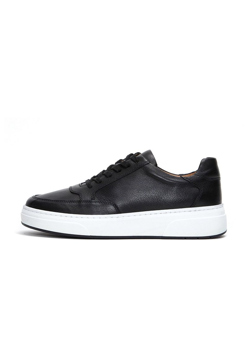 Zapatilla de cuero negro con punta redondeada, suela de goma blanca y seis agujeros para los cordones. Presenta un upper texturizado y detalles de costura en contraste.