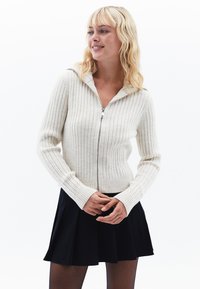 Jasna kremowa kardigan o żeberkowanej fakturze z zamkiem z przodu, długimi rękawami i dopasowanym krojem. Noszona na czarną rozkloszowaną spódnicę.