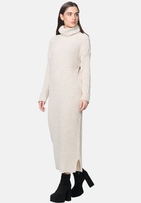 Cremefarbenes, geripptes Strickkleid mit hohem Rollkragen, langen Ärmeln und seitlichen Schlitzen; kombiniert mit schwarzen Plateau-Stiefeletten.