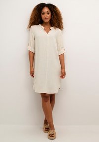 Abito tunica beige chiaro con colletto, maniche arrotolabili e spacchi laterali, realizzato in tessuto testurizzato, abbinato a sandali slip-on intrecciati.