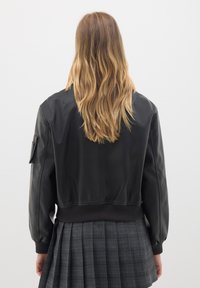 Giubbotto bomber in pelle nera con polsini e orlo a costine, visto da dietro, abbinato a una gonna a pieghe in plaid grigio.