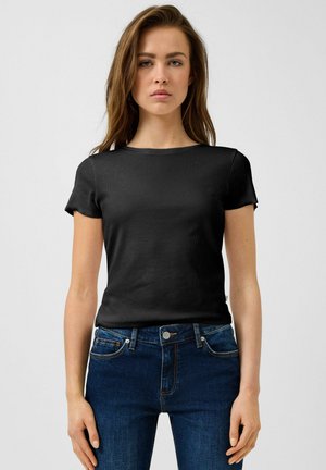 Junge Frau trägt ein eng anliegendes schwarzes Kurzarmshirt und blaue Jeans, steht vor einem schlichten hellgrauen Hintergrund.