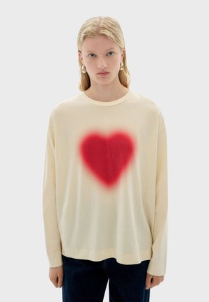 Crème kleurige longsleeve shirt met een grote, rode hartgraphic in het midden, gekenmerkt door een zachte textuur en een ontspannen fit, gecombineerd met donkere denim broek.
